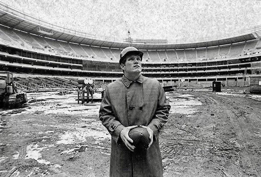 A person wearing a coat and hat stands in the middle of a large, partially constructed or renovated stadium. They are holding a football and looking upwards. The ground is muddy, and construction equipment is visible in the background.