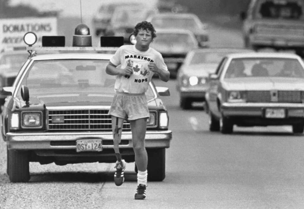 A man wearing a "Marathon of Hope" T-shirt runs on a road, followed by a police car and several vehicles. The road has a sign displaying "Donations." The scene captures a determined expression and supportive atmosphere.