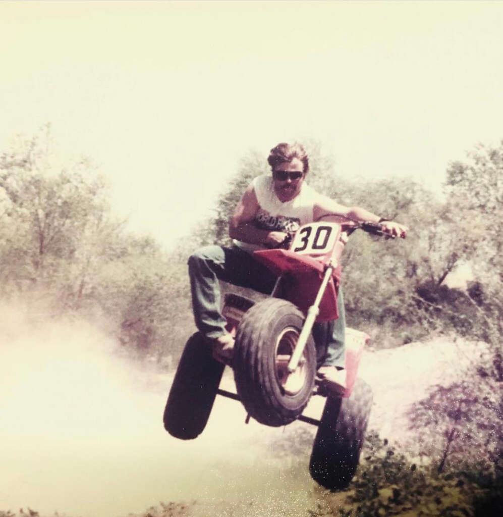 Person riding a red three-wheeled ATV with the number 30, airborne on a dirt trail. The rider wears sunglasses and a sleeveless shirt, with trees and dust visible in the background.