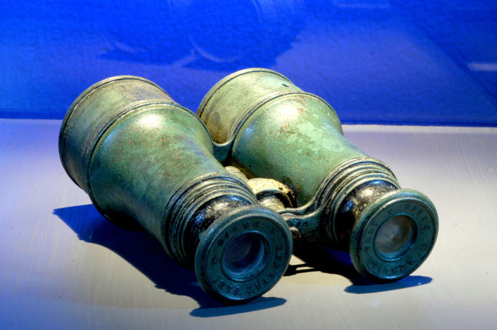 Antique binoculars with a weathered, greenish patina rest on a reflective surface, illuminated by a blue background. The eyepieces display engraved text, adding to their vintage appearance.