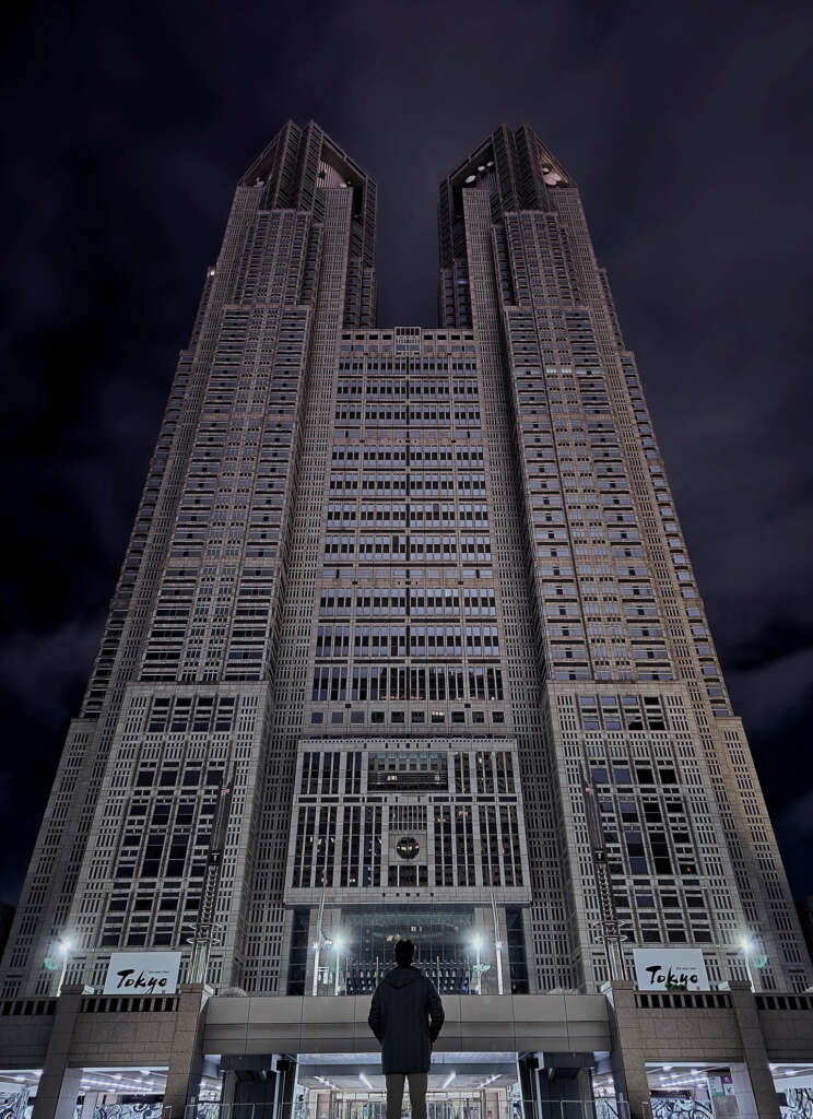 A person stands in front of a tall, illuminated skyscraper at night, looking up. The building has intricate architectural details with numerous windows, and the sky is dark and cloudy.