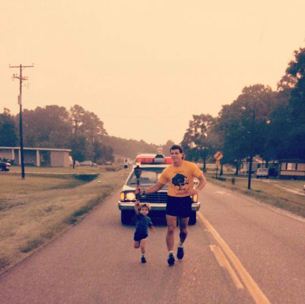 A man and a young child jog in front of a police car with flashing lights on a suburban street lined with trees and houses. The man wears a yellow shirt and dark shorts, while the child holds his hand and wears a blue outfit.