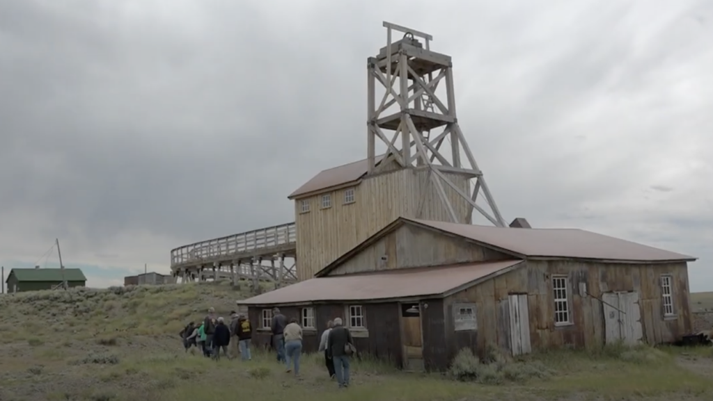 A group of people walking toward a rustic wooden mining structure in a grassy, open area. The sky is cloudy, and there are other small buildings in the background. The scene conveys a historical or abandoned setting.