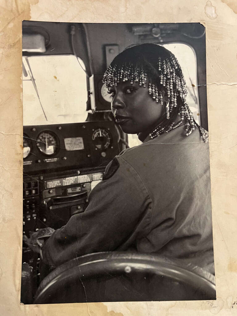 Black and white photo of a person with beaded hair sitting in the cab of a truck, facing slightly to the side with hands on the wheel. The dashboard with gauges is visible in the background. The image has a vintage and worn appearance.