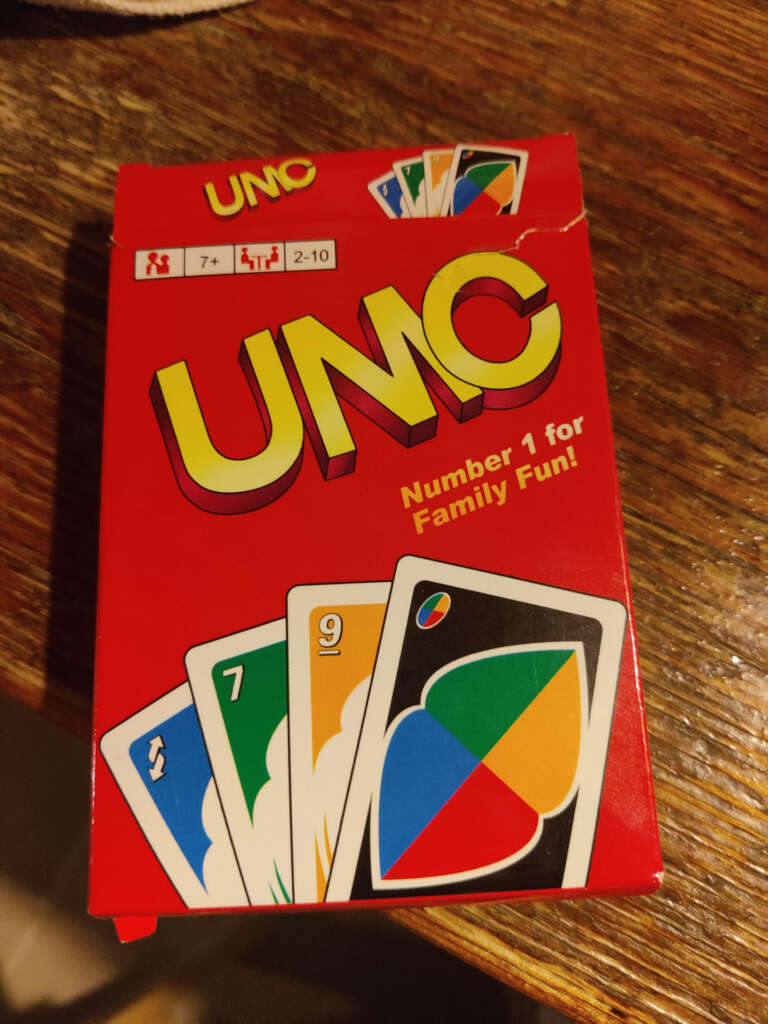 A red UNO card game box is on a wooden surface. The box displays the words "Number 1 for Family Fun!" and shows images of colorful playing cards. It is suitable for ages 7+ and can be played by 2-10 players.