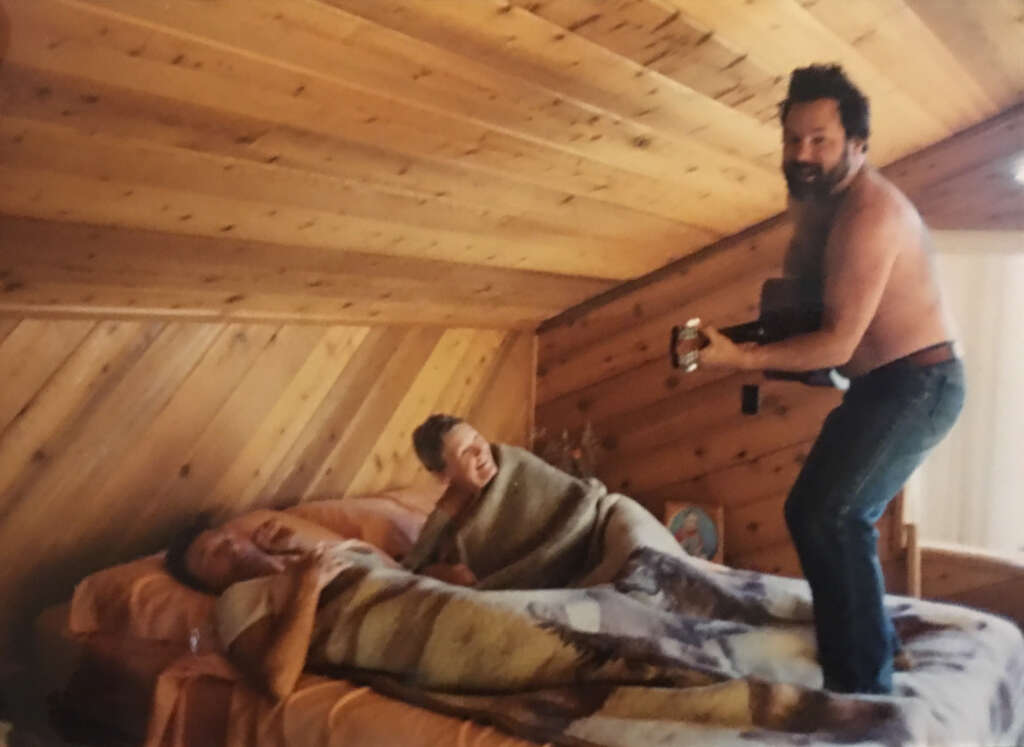 A man with a beard, shirtless, plays a guitar standing on a bed. Two people lie on the bed under a blanket, laughing. The room has wooden paneled walls and ceiling, creating a cozy cabin-like atmosphere.