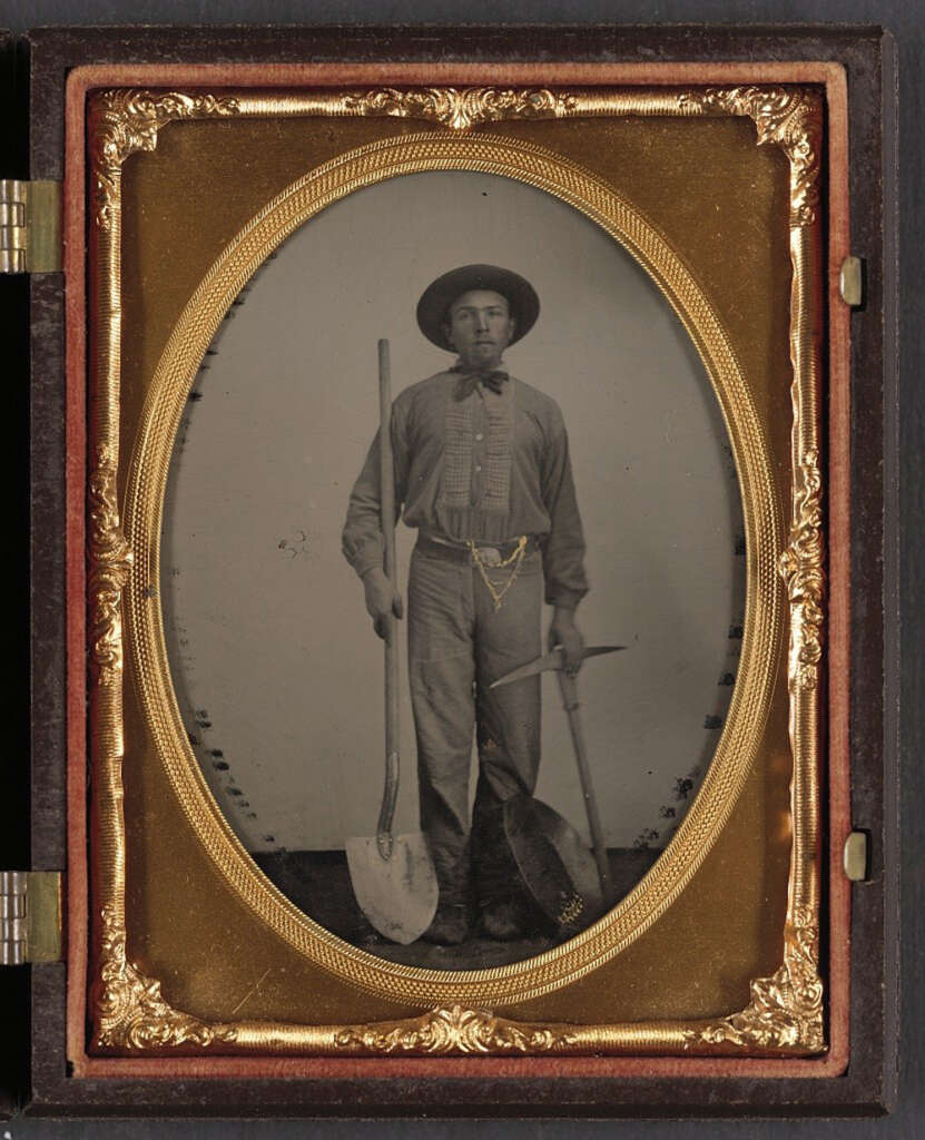 A sepia-toned portrait of a man in 19th-century work attire holding a shovel. He wears a hat and stands in front of a neutral backdrop. The ornate frame has gold detailing, enhancing the vintage appearance.