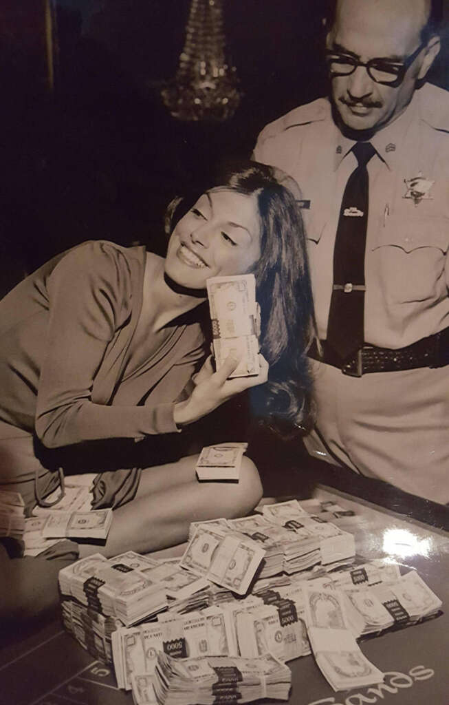 A woman smiles joyfully, holding a stack of cash to her face, while a man in a sheriff's uniform stands beside her. They are surrounded by a table piled with stacks of money.