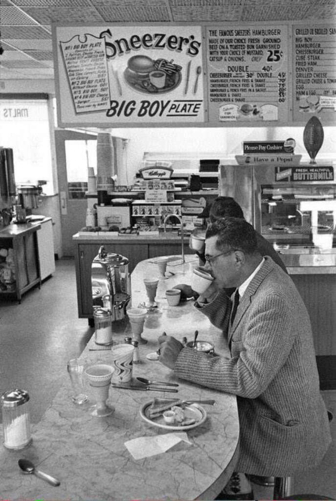 Black-and-white photo of a vintage diner counter. Two men in suits sit on stools, eating. A sign overhead reads "Neezer's Big Boy Plate" and lists prices. The counter holds dishes, napkin dispensers, and utensils.