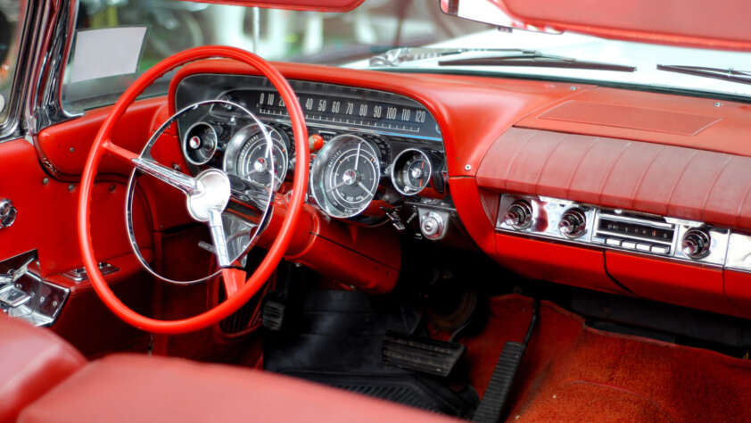 Interior of a vintage car featuring a red leather dashboard and steering wheel. The dashboard includes classic analog gauges and a radio. The car’s upholstery and carpet are also in matching red.