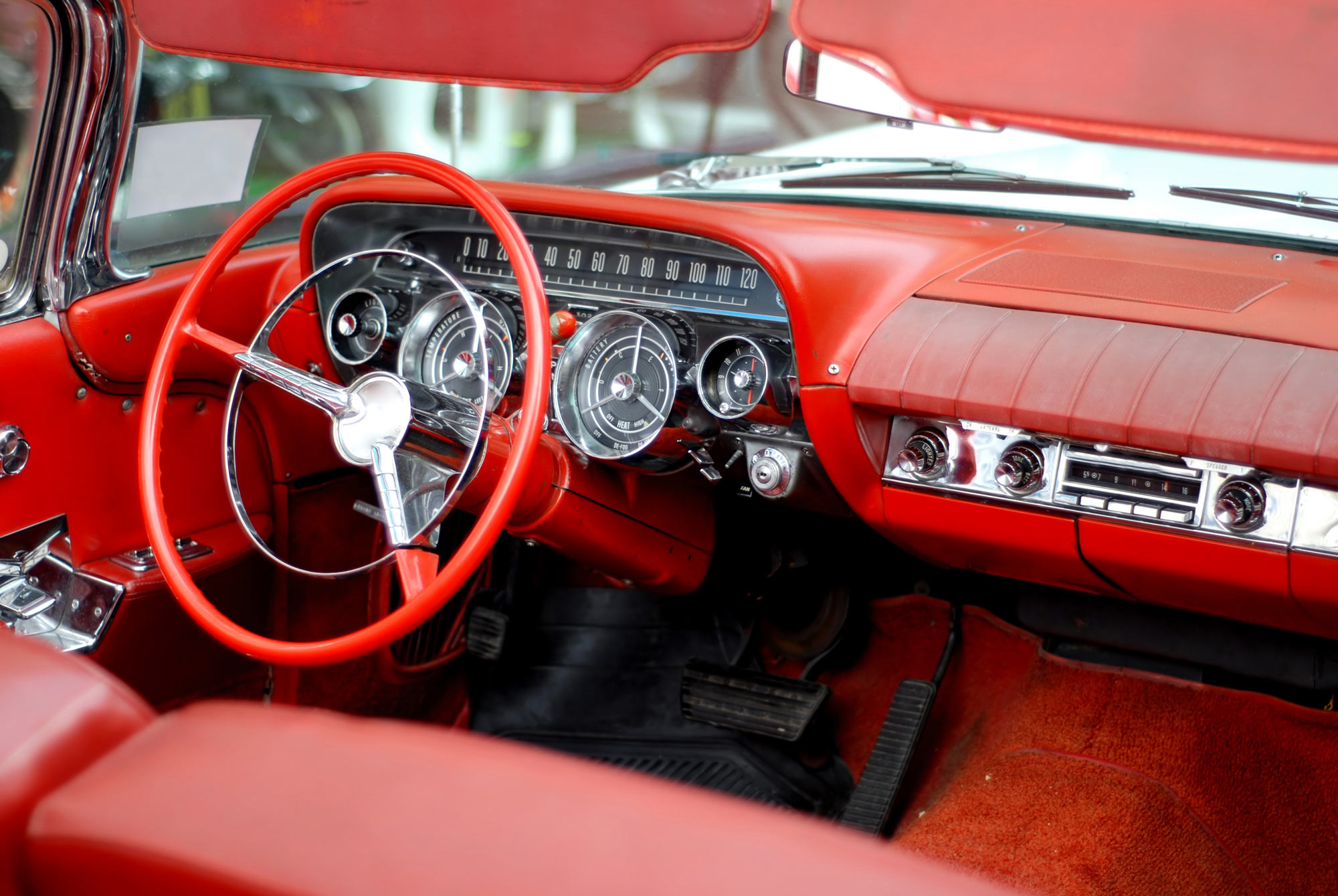 Interior of a vintage car featuring a red leather dashboard and steering wheel. The dashboard includes classic analog gauges and a radio. The car’s upholstery and carpet are also in matching red.