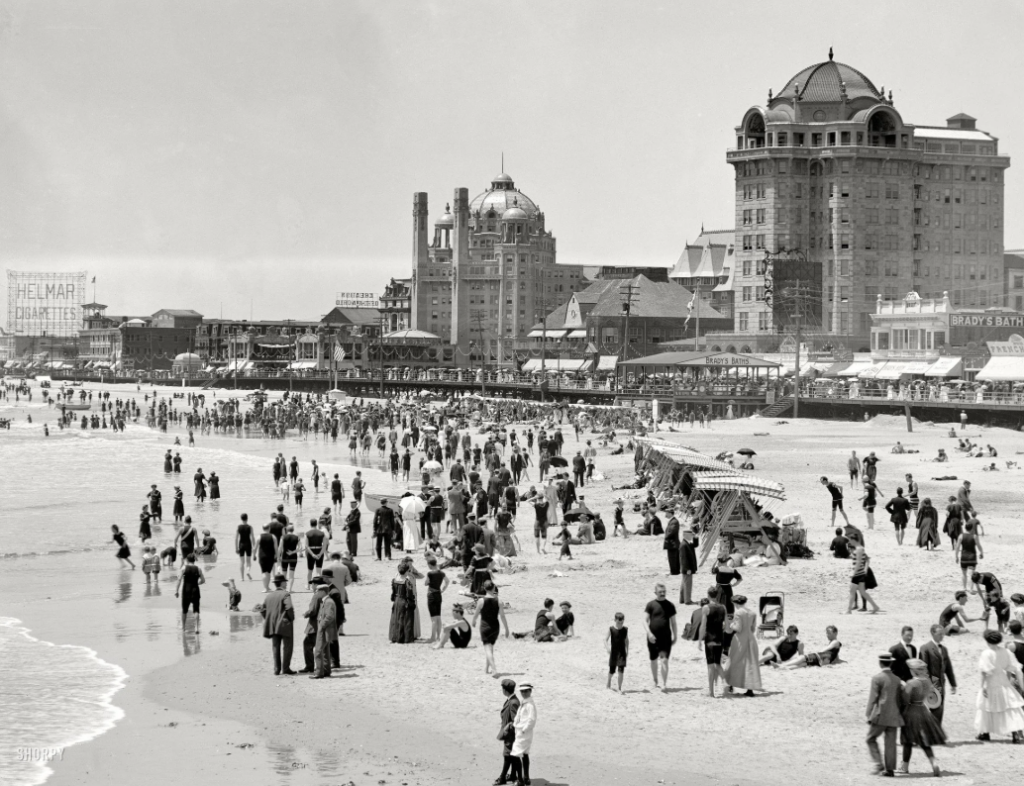 A black and white photo of a crowded beach scene from the early 1900s. People are dressed in period swimwear and formal clothing. Large buildings and a boardwalk are visible in the background, and umbrellas dot the sandy shore.