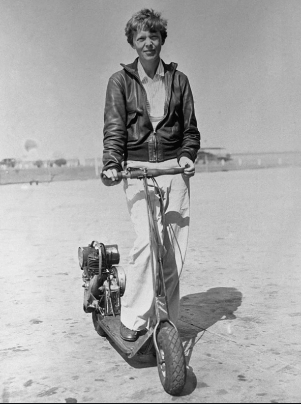 A person in a leather jacket and light pants stands on a motorized scooter on a beach. The background includes a clear sky and blurred structures or vehicles in the distance.