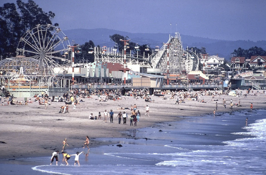 A bustling beach scene with people enjoying the sand and waves. In the background, there is an amusement park with a Ferris wheel, roller coaster, and other attractions. The sky is clear, and the ocean waves gently lap the shore.