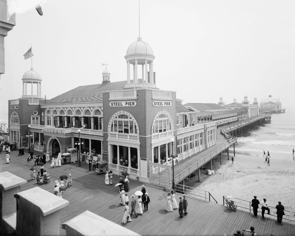 A historic black-and-white photo of Steel Pier, featuring a grand building with arches and towers. People in early 20th-century clothing stroll along the boardwalk, with the ocean and sandy beach visible in the background.