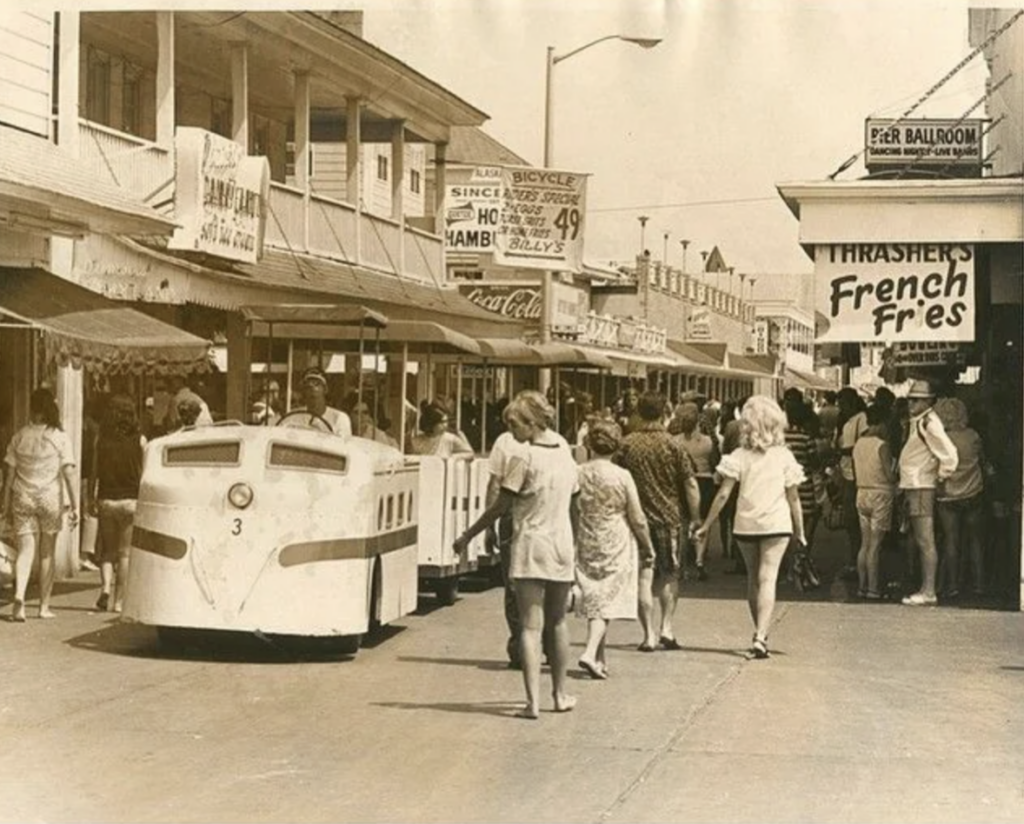 Vintage street scene with people walking and a small white train driving down the road. Storefronts display various signs, including one for Thrasher's French Fries. The atmosphere is lively and bustling.