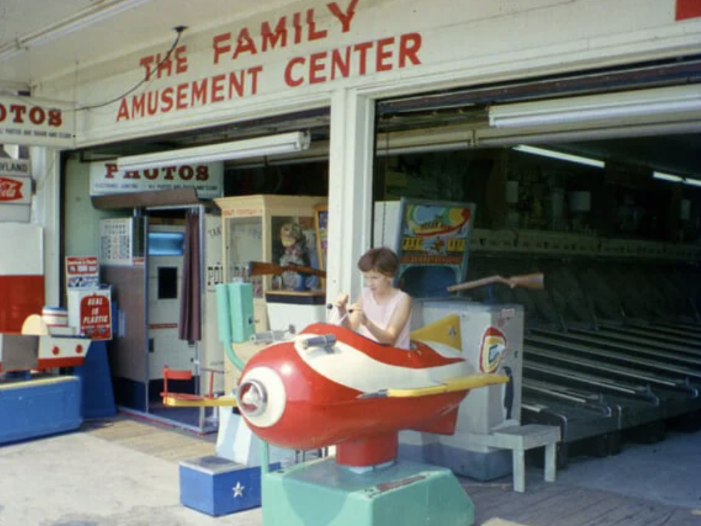 A young girl sits in a red and white coin-operated airplane ride outside "The Family Amusement Center." The background shows arcade machines and a photo booth. The scene has a nostalgic, retro vibe.