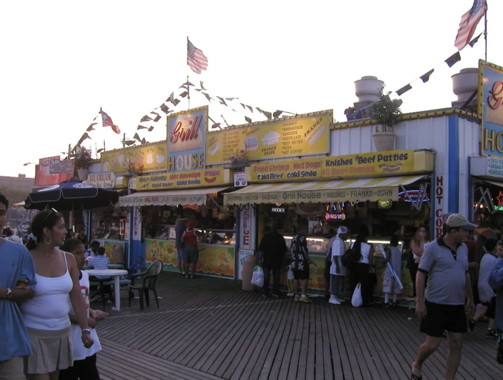 People are walking past a bustling boardwalk food stand with colorful signs advertising items like hot dogs and soda. Flags and banners decorate the building. The scene captures a lively, casual outdoor atmosphere.