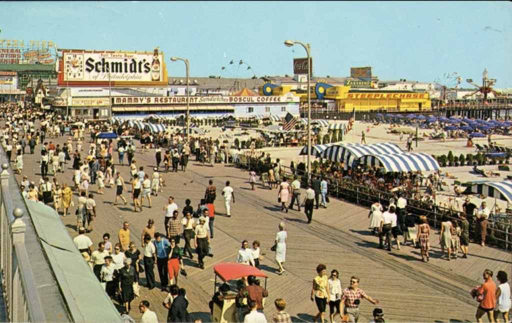 A crowded boardwalk scene with people walking along the wooden planks under a clear sky. Numerous signs for Schmidt's, Danny's Restaurant, and Boshul Coffee are visible. The beach and amusement park rides are in the background.