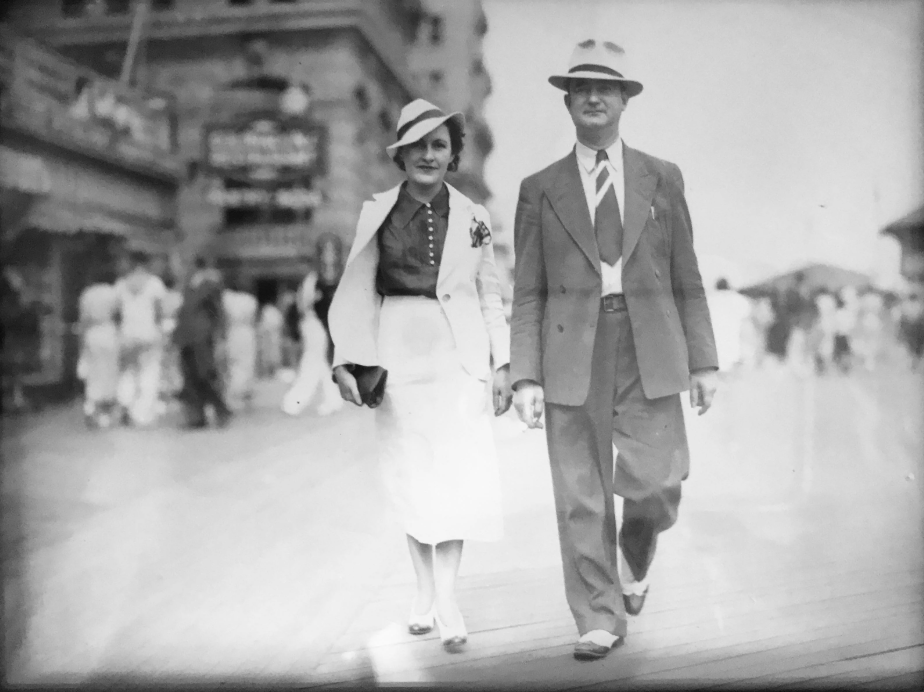 A black and white photo showing a couple walking confidently on a boardwalk. They are dressed in vintage 1930s style, with the man in a suit and fedora and the woman in a tailored dress with a hat. The background shows a blurred crowd and buildings.