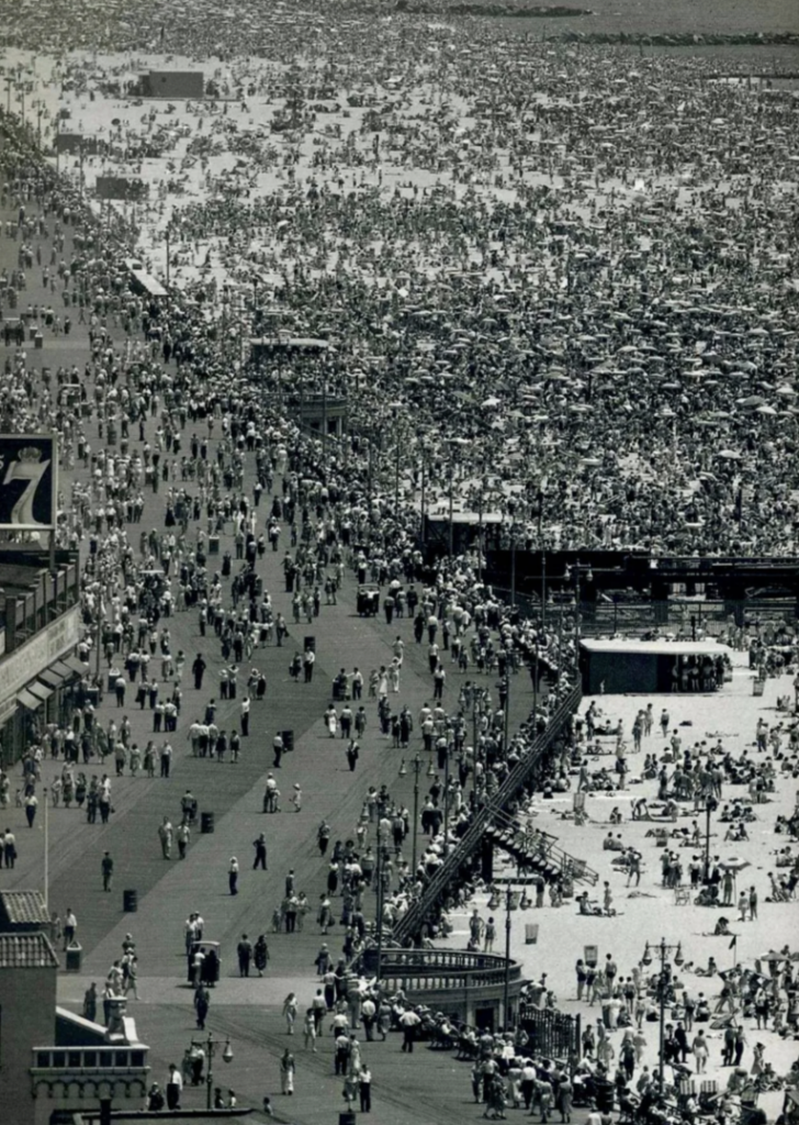 Aerial view of a crowded beach and boardwalk. Thousands of people are gathered, with many sunbathing and relaxing on the sand. The boardwalk is filled with people walking. The ocean is visible in the background. The scene is in black and white.