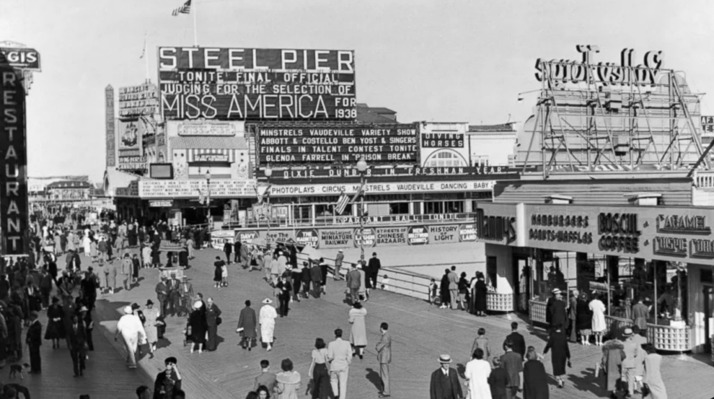 A bustling 1938 boardwalk scene with people walking near signage for Steel Pier, advertising events like Miss America judging and various performances. Shops and attractions are visible, including a famous soda sign and a restaurant.