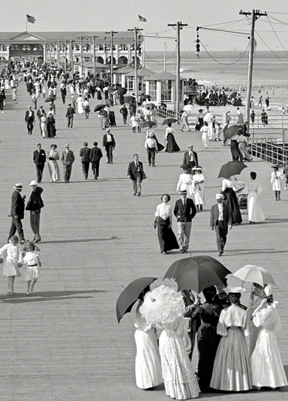 Black and white photo of a crowded boardwalk by the beach. Men, women, and children are dressed in early 20th-century clothing. A group of women gathers with umbrellas, while others stroll along the wooden path. Telephones poles and buildings are visible.