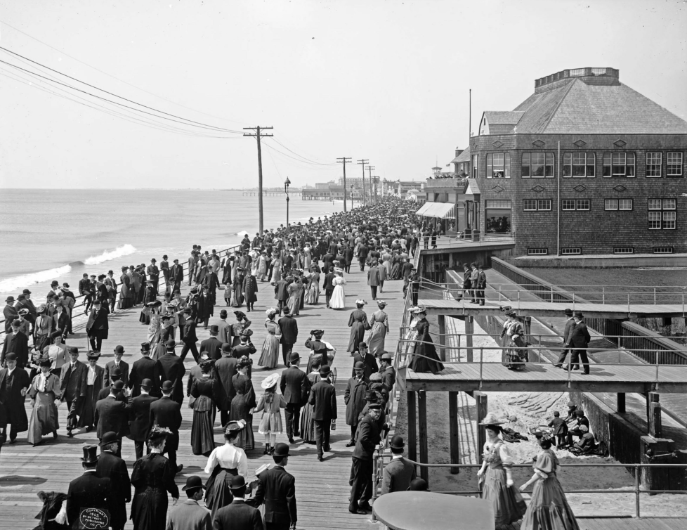 Crowded boardwalk scene by the beach, featuring people in early 20th-century attire, some walking, others pausing to chat. A large brick building is on the right, with telephone poles running alongside the ocean on the left.