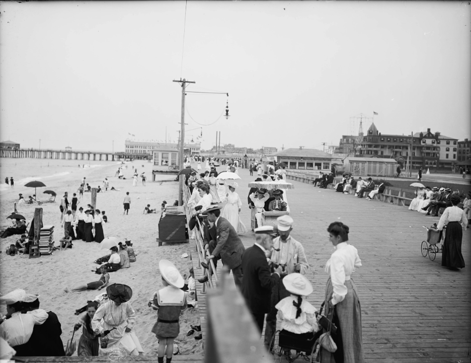 Black and white photo of a crowded beach boardwalk in the early 1900s. People in period clothing stroll, sit, and gather along the boardwalk, with a beach on the left and ocean piers in the distance.