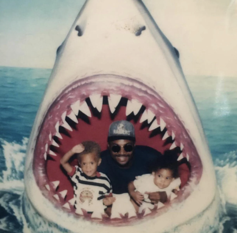 A man in sunglasses and a hat poses with two small children inside a large shark mouth prop by the ocean. The children smile, one with their hand raised. The scene is bright and playful, creating a humorous impression of being inside a shark's mouth.