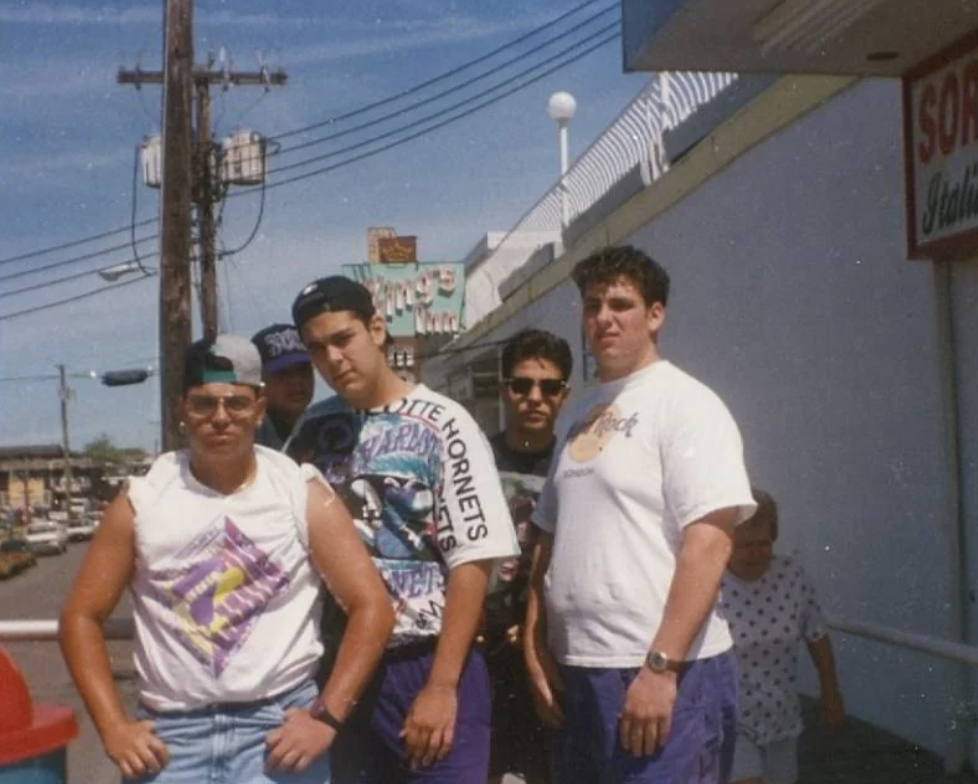 Five young men stand on a sidewalk in casual 90s attire, including T-shirts and baseball caps. They pose confidently with a sunny street and store signs in the background.