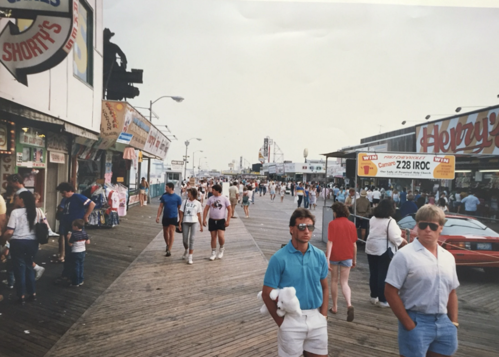 People are walking along a busy boardwalk with shops on either side, including a candy shop and a games arcade. A man holds a stuffed toy while others are engaged in various activities, creating a lively seaside atmosphere.