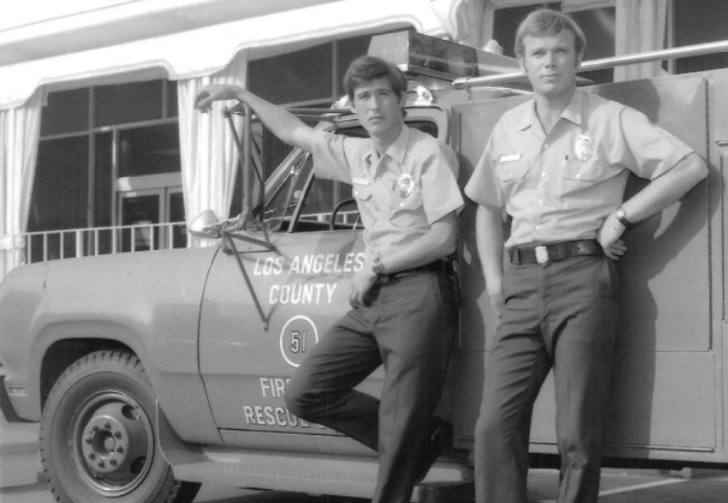 Two men in uniform stand in front of a Los Angeles County Fire Department rescue vehicle. One leans against the truck with his arm, while the other stands with hands on hips. The background shows a building with large windows and curtains.