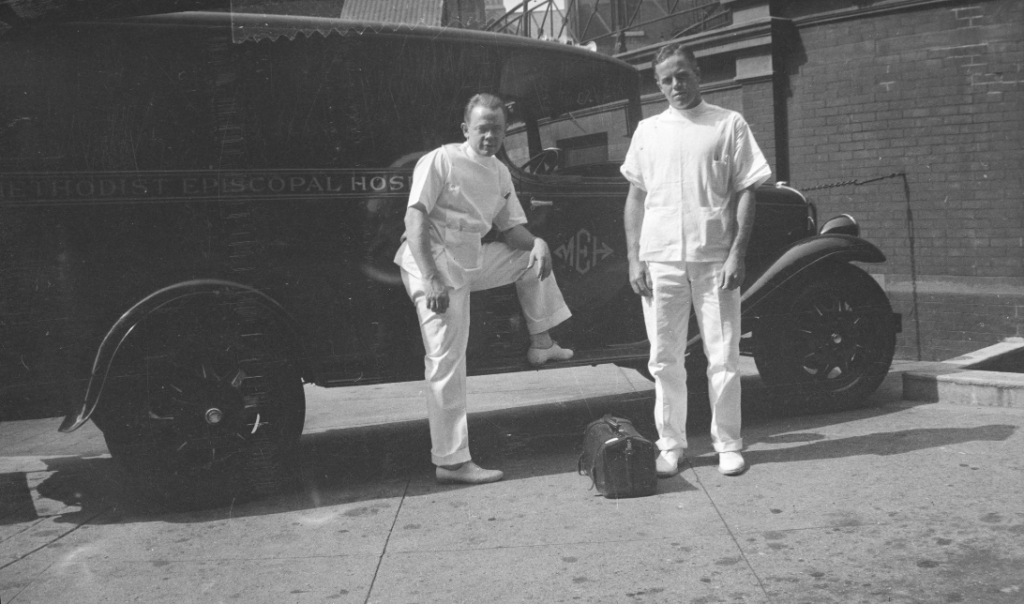 Two men in white uniforms stand beside an early 20th-century ambulance with "Methodist Episcopal Hospital" written on it. One man leans on the vehicle, and a medical bag is on the ground. They're outside a brick building.