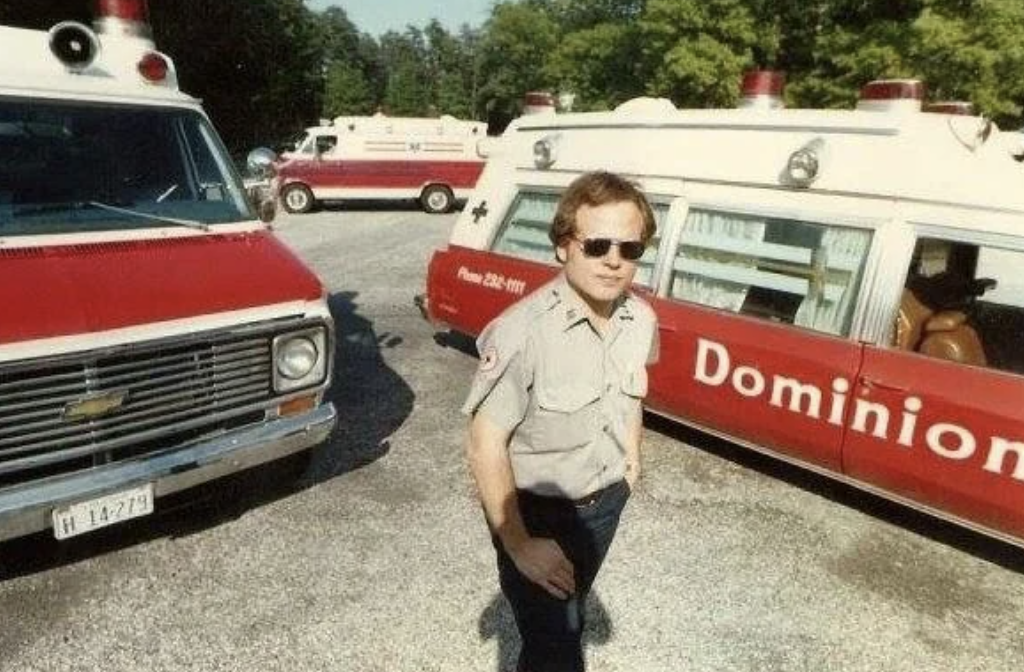 A man in a uniform stands outdoors in front of several red and white ambulances parked on a gravel surface. The ambulances have the word "Dominion" visible on the side. Trees are in the background.