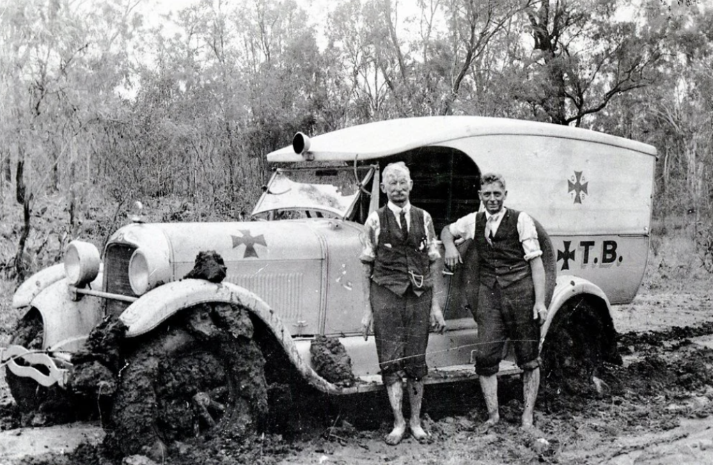 Black-and-white photo of two men standing beside a vintage, mud-covered vehicle with a cross symbol and "T.B." letters on the side. Both are wearing vests, ties, and rolled-up sleeves. The background features trees and brush.