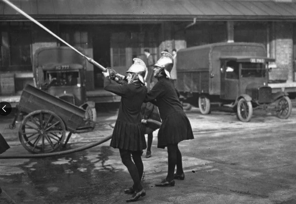 Two women in vintage firefighter uniforms aim a hose upwards, practicing outside with water spraying. Old trucks and a wagon are in the background, suggesting an early 20th-century setting.