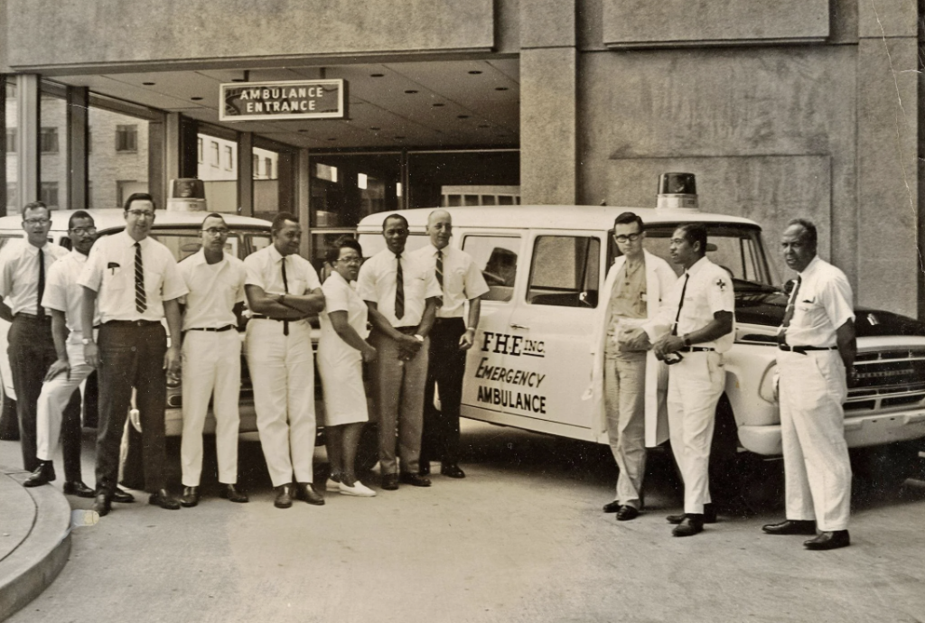 A group of men and women stand in front of an emergency ambulance parked at a hospital entrance. They are wearing formal attire, and some have lab coats. A sign above reads "Ambulance Entrance." The group appears to be hospital staff.