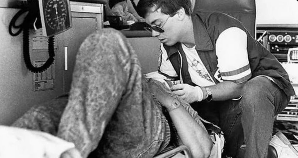 A paramedic wearing sunglasses attends to a patient inside an ambulance. The patient is lying down while the paramedic kneels beside them, performing a medical procedure or check-up. Medical equipment is visible in the background.