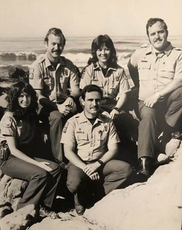 A vintage black and white photo of five park rangers in uniform posing on rocky coastal terrain. Three are seated on rocks, while two stand behind them. The ocean is visible in the background, with waves crashing on the shore.