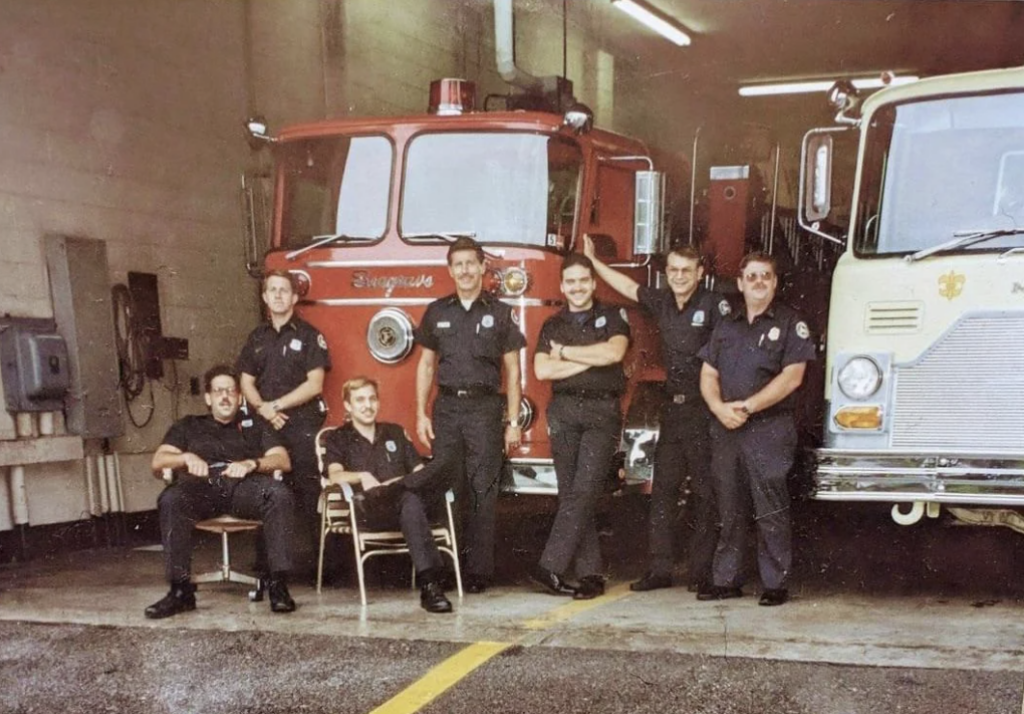 Seven firefighters in uniform stand and sit in front of two fire trucks inside a garage. One firefighter sits on a chair, and the others stand or lean casually. The trucks are parked side by side, with one red and one white.