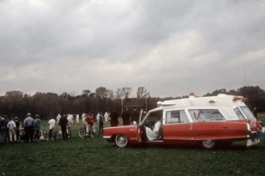 A red and white vintage ambulance is parked on a grassy field under a cloudy sky. A group of people, some on bicycles, gather nearby, watching a sports event in the distance.