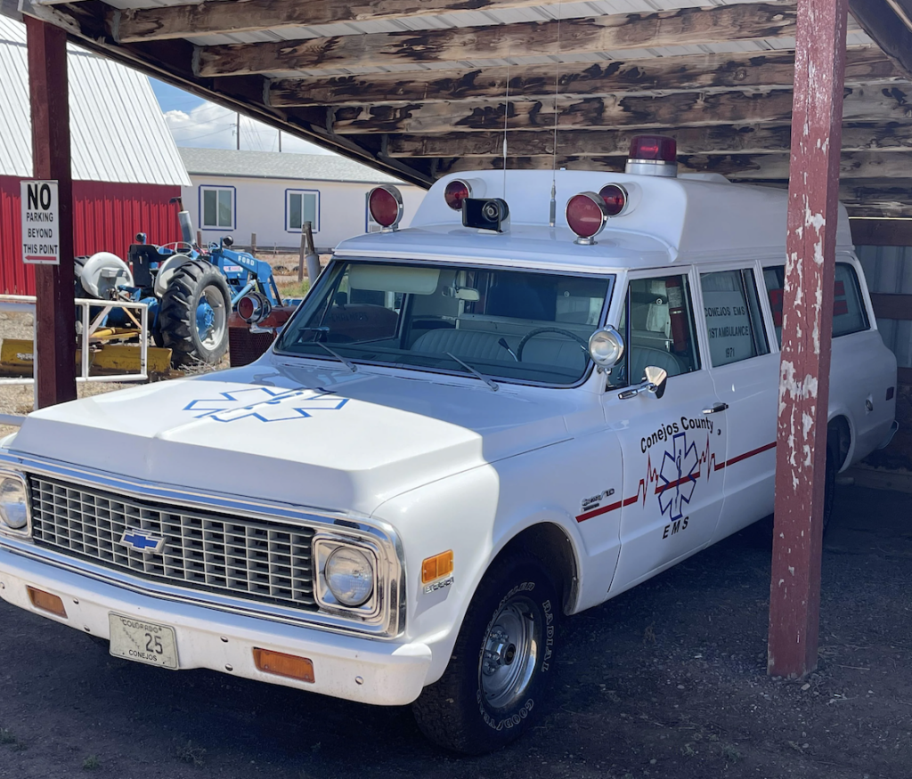 A vintage white EMS vehicle with a red medical symbol and "Conejos County EMS Ambulance" text is parked under a wooden shelter. It has a classic Chevrolet front, and red lights on top. A blue tractor and red building are visible in the background.