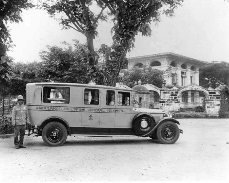 A vintage ambulance with "Philippine General Hospital" written on the side is parked on a road. A man in a military-style uniform stands beside it. There's a large historic building and lush trees in the background.