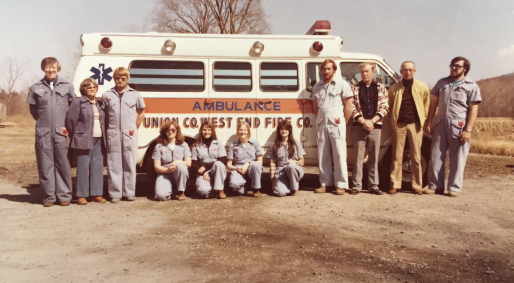 A group of people in uniform pose in front of a vintage ambulance labeled "Union Co. West End Fire Co." There are nine individuals standing and two kneeling, with a rural background and clear sky.