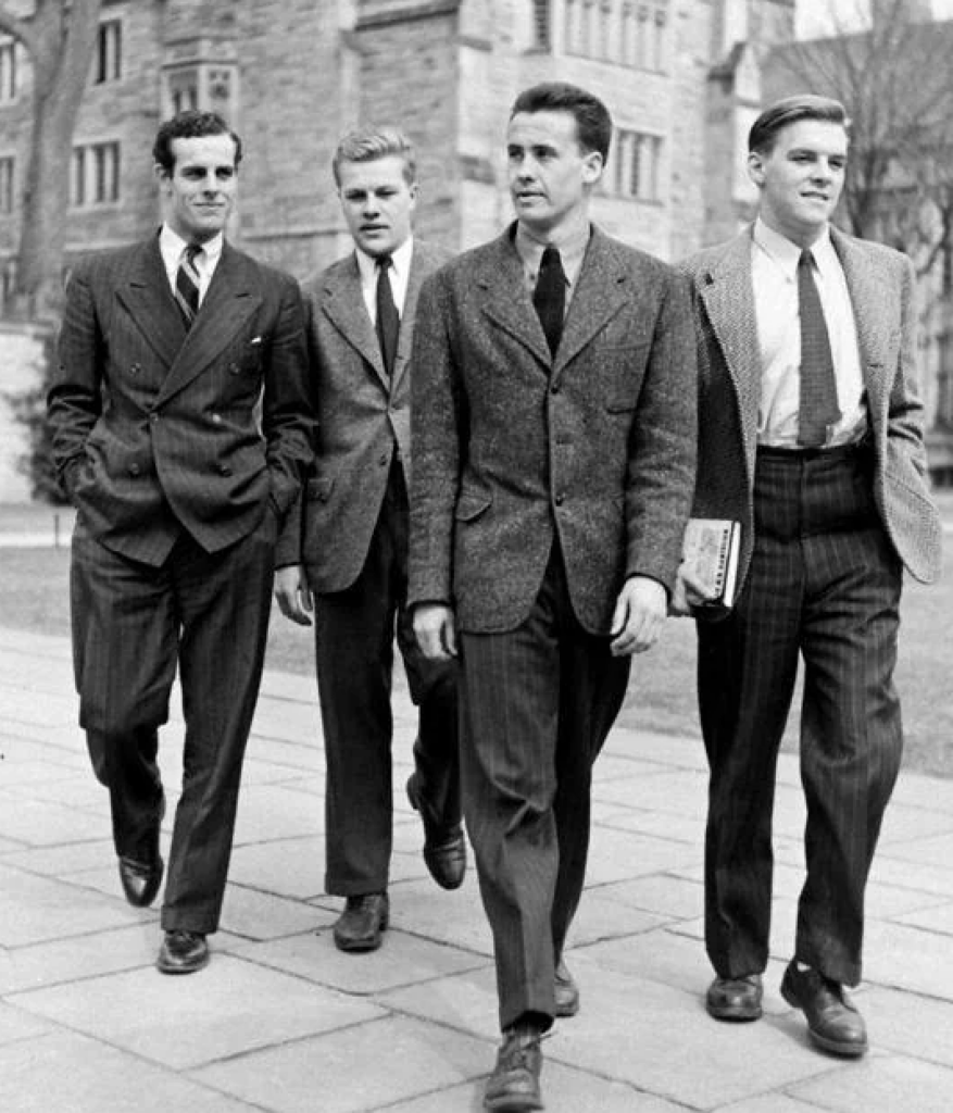 Four men in suits walk together on a paved path in an outdoor setting with buildings in the background. One man holds a book under his arm. They appear to be engaged in conversation, dressed in mid-20th century style.