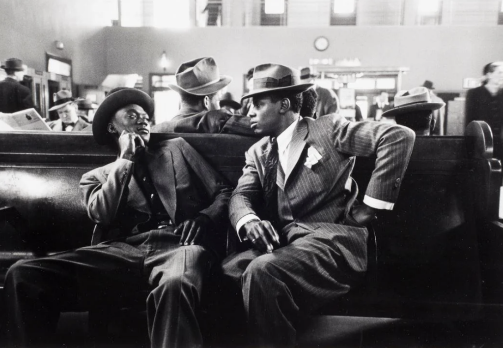 Two men in suits and fedoras sit on a bench in a busy train station, engaging in conversation. One man leans back with his arm across the bench, while others in the background read newspapers or chat. The scene captures a vintage atmosphere.