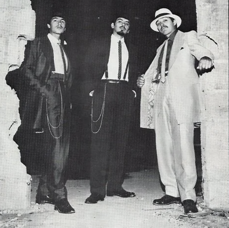 Three men stand confidently in a worn archway, wearing 1940s-style zoot suits. One wears a light suit with a hat, and the other two wear dark suits. Each has a distinctive pose, conveying an air of swagger and style.