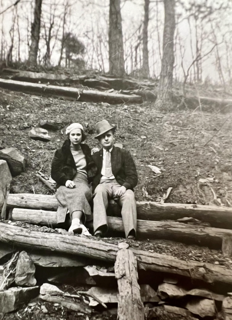 A black and white photo of a man and woman sitting on wooden logs in a forest. The woman wears a coat, skirt, and headscarf, and the man wears a suit with a bow tie and hat. Bare trees surround them on the slope.