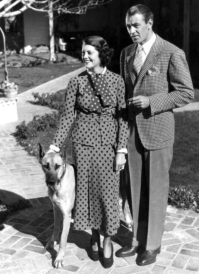 A couple in vintage clothing stands outdoors on a stone path, accompanied by a large dog. The woman wears a polka dot outfit, and the man is in a checkered blazer. They are in a sunny garden setting.